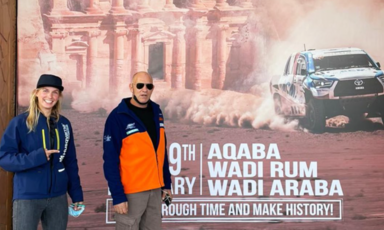 A man and a woman pose in front of a large poster featuring a racing car 