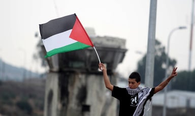A Palestinian boy wearing a kuffiyeh holds a Palestinian flag in front of a military watch tower