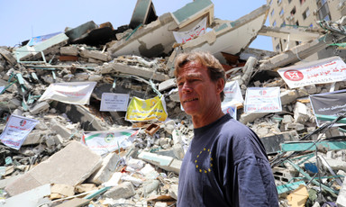 Man wearing T-shirt stands in front of destroyed building 