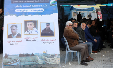 Men sit on plastic chairs under a large tent next to a banner featuring pictures of three men 
