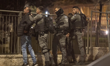 A police officer, accompanied by other officers carrying weapons, grabs a civilian from behind. 