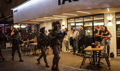 People stand in front of restaurant as Israeli soldiers walk along street