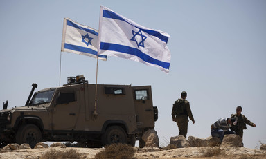 Two blue and white flags fly beside an armored vehicle and soldiers in uniform 