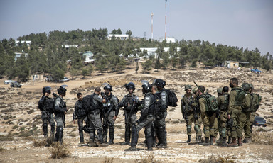 Soldiers stand on bare ground in front of forested hilltop settlement