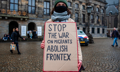 Woman wearing mask holds a placard reading Stop the war on migrants abolish Frontex