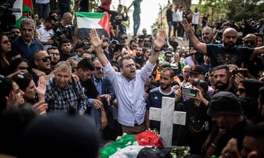 Man holds his hands above his head while standing in crowd around grave with Palestine flag on it