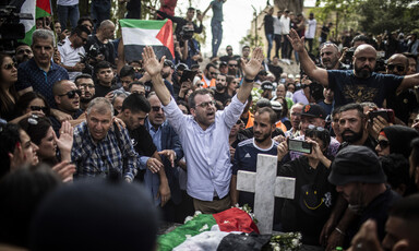 Man holds his hands above his head while standing in crowd around grave with Palestine flag on it