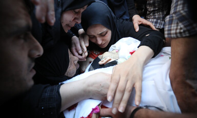 Women clasp hands and cry while leaning over shrouded body