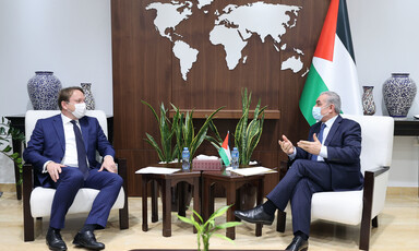 Two men wearing suits face each other while sitting in chairs with a table in between them