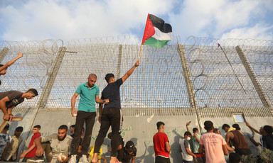 A youth holds a Palestine flag in front of a tall militarized fence