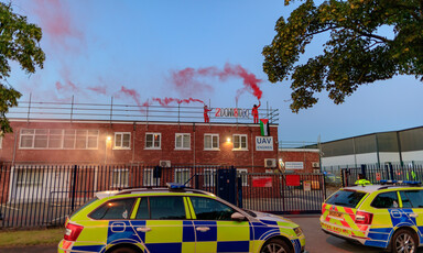 Two people stand on top of a building, with red smoke above them