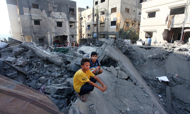 Two boys sit on ruins of fallen building while surrounded by bombed-out midrise buildings