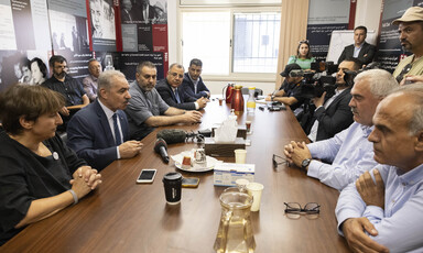 Around a dozen people sit at and stand around conference table