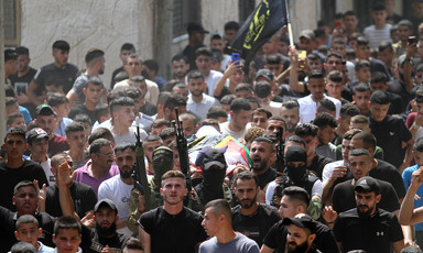 Men carry a body on a stretcher in a crowd that includes some with rifles 