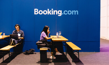 Two people sit on benches and work in front of a Booking.com wall 
