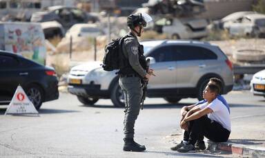 An Israeli soldier stands in front of two youths sitting on curb at checkpoint