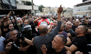 A flag drapes a body on a stretcher carried by a large crowd