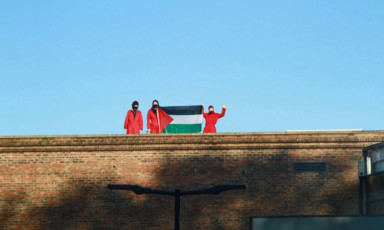 Three people in red hold up a Palestinian flag on a rooftop