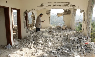 Man holding mobile phone stands in rubble of destroyed home with holes in walls showing view of hill