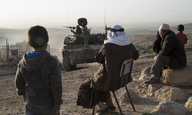 Young children stand and older men sit along a dirt road with an Israeli tank approaching