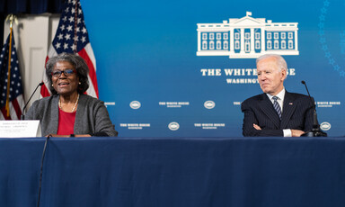 Two people sit at a table with American flags behind them