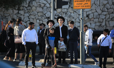 Boys in Orthodox dress and a boy with a scooter stand at a traffic light as other people walk by
