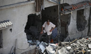 A man looking down with his arms akimbo stands on rubble in a hole in the wall of a bombed-out building