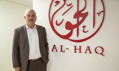 Shawan Jabarin, wearing sports coat and collared shirt, stands in front of wall bearing a large reproduction of Al-Haq's calligraphic logo