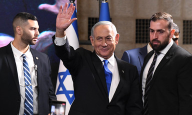 Benjamin Netanyahu, wearing suit and tie, waves while standing in front of Israeli flags