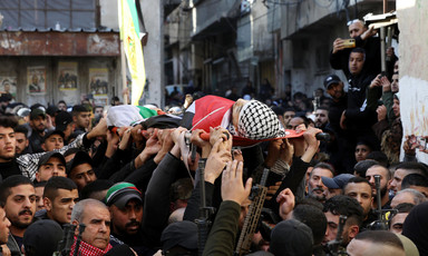 Crowd of men, some carrying rifles, stand in street while body shrouded in Palestine flag is carried on stretcher