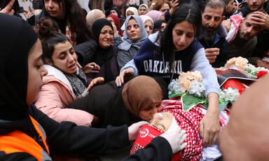 Women and girls cry while embracing and kissing boy shrouded in red kuffiyeh and covered with flowers