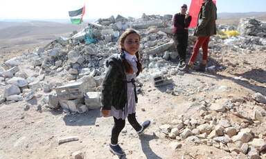 Girl in school uniform stands in front of rubble with Palestinian flag flying over it as two other people stand behind