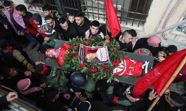 Men and boys carry the body of a boy wrapped in a flag and covered in a wreath and a poster on a stretcher 