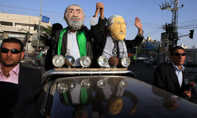 Two men wearing masks depicting Ismail Haniyeh and Mahmoud Abbas wave from a black car