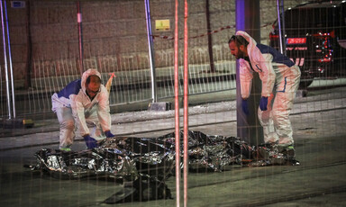 Two men in white hazmat suits stand over bodies wrapped in reflective material