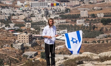 Young man in religious dress holds Israeli flag while standing in front of landscape with Palestinian houses