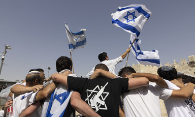 Young men seen with their backs to camera embrace with Israeli flags waving above them. One is wearing a black T-shirt showing the Star of David with a rifle in the middle.