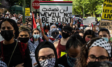 A group of protesters hold signs and Palestine flags. One banner reads We Will Not Be Silent in English and Arabic.