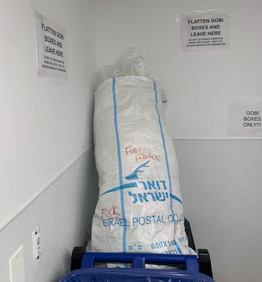 A discarded Israeli postal service bag sits in a recycling bin. Fuck is written above the word Israel, and Free Palestine is written on the top in red marker. 