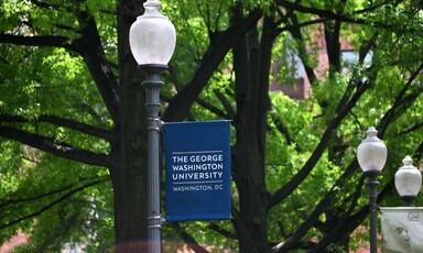 A blue banner with the name of the university hangs on a black lamppost, with trees in the background 