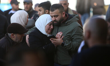 Man looking down and holding mobile phone wraps arm around crying woman as other people stand around them