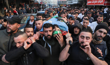 Crowd of men in street carry shrouded body on stretcher