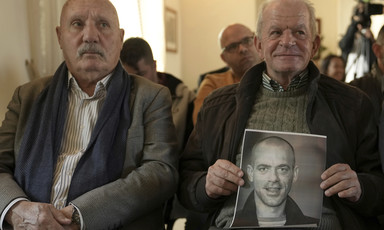 Two elder men sit next to one another and one holds a photo of a younger man