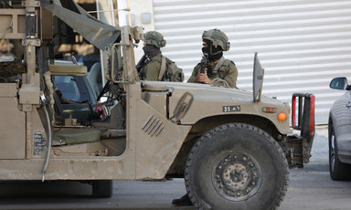 Two soldiers stand near armored vehicle, one of them pointing a sniper 