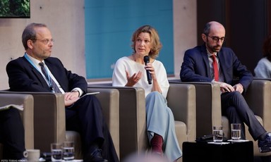 Katharina von Schnurbein speaks into a microphone and gestures with her hand while sitting in between two men on stage