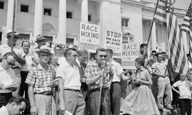 Man speaking at microphone in front of crowd at the Arkansas State Capitol protesting school integration with signs reading Race mixing is Communism and Stop the race mixing