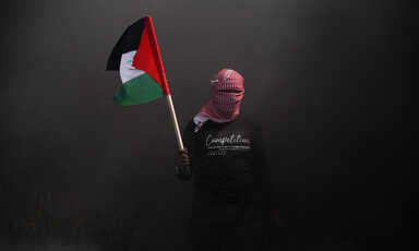 A person with their face covered emerges through dark smoke with the Palestinian flag