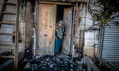 A women peers out from a metal door to an area covered in burned debris