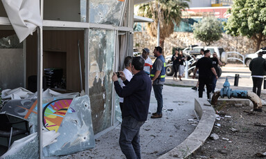 Men stand in front of building with broken and shrapnel-pocked windows