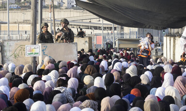 Two Israeli armed soldiers stand behind concrete blocks above dozens of Palestinians standing at checkpoint